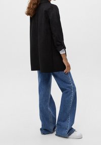 Long blazer en suède noir avec col, manches retroussées, associé à un jean en denim bleu à jambes larges et des baskets blanches. Texture lisse, coupe décontractée.