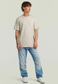 Beige t-shirt med kort ärm och avslappnad passform, tillsammans med ljusblå jeans med lappar och vita sneakers. Enkel design utan framträdande mönster.