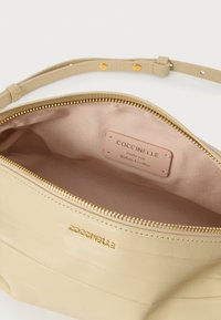 Beežist nahast käekott kuldse tõmblukuga, sileda tekstuuriga ja heleda kangase voodriga. Sisemine silt kannab kirjapilti "Coccinelle Autentne Itaalia Nahk".