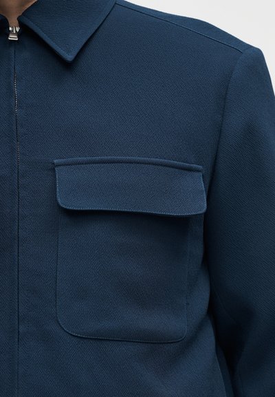 Tmavomodrá textúrovaná bunda s plochým golierom, náprsníkovým vreckom s klopou a predným zapínaním na zips.