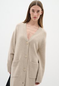 Beige stickad cardigan med en djup V-ringning, fem knappar, ribbade ärmmuddar och två framfickor. Tillverkad av mjukt, strukturerat tyg.