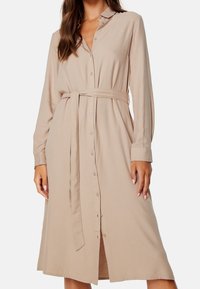 Beżowa sukienka zapinana na guziki z długimi rękawami, kołnierzykiem i pasującym paskiem. Wykonana z gładkiego materiału, ma średniej długości dół i uniwersalny krój.