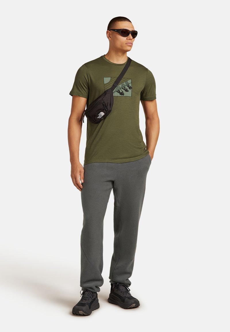 Camiseta verde oliva con un diseño gráfico, combinada con pantalones de chándal grises y zapatillas negras. El accesorio incluye un bolso cruzado negro.