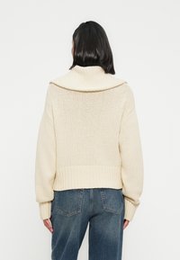 Πλεκτό πουλόβερ σε κρεμ χρώμα με γιακά τύπου χωνιού, ριμπ τελείωμα και μακριά μανίκια. Υφή υφάσματος, άνετη εφαρμογή και cropped μήκος.