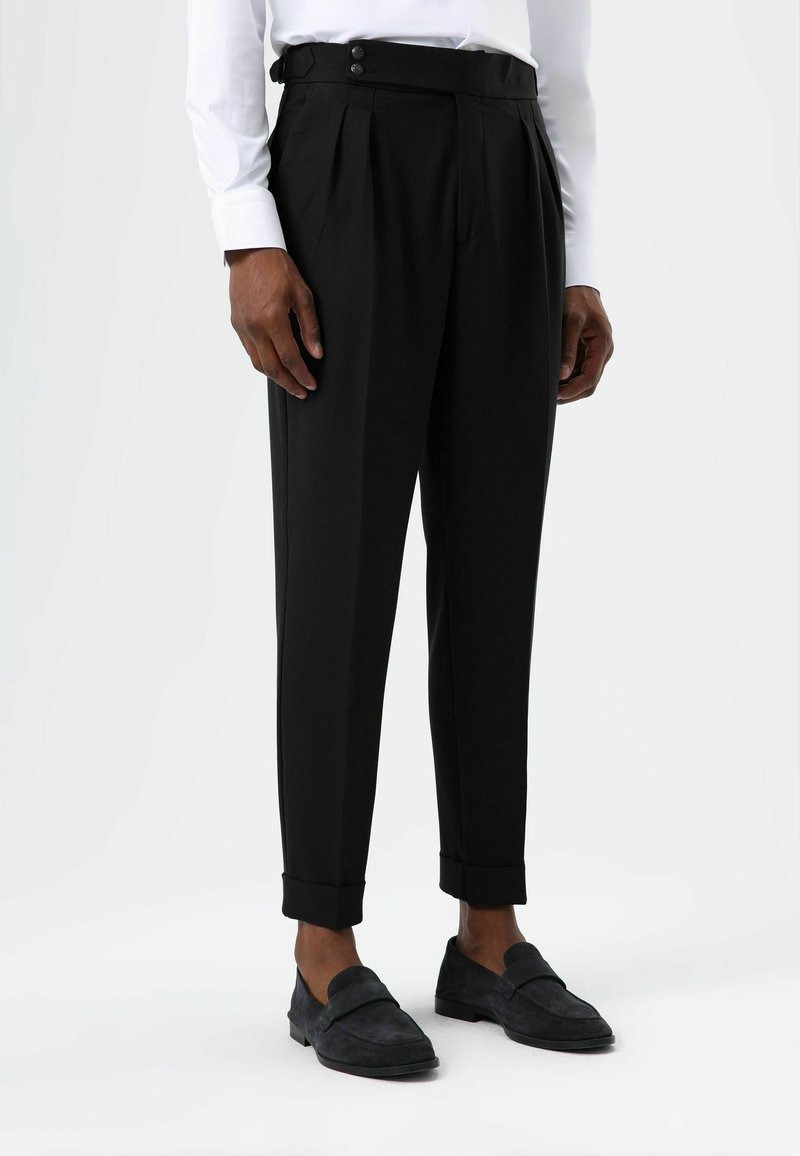Pantalon noir plissé avec une coupe ajustée, ourlets retroussés et une taille haute ornée de deux boutons noirs. Porté avec des mocassins en daim foncé.
