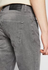 Sivevaa jeans iz naravi gladke teksture, pet žepov in črna usnjena paketa z blagovno znamko na hrbtni strani. Prileganje je ozko z blagimi znaki obrabe.