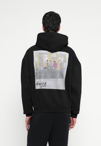 Fekete kapucnis pulóver nagy fényképes mintával a hátoldalán, utcai jelenettel és alakokkal. Puha anyag, kényelmes szabás, kenguruzseb.