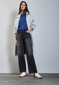 Grauer Mantel über blauem Oberteil, schwarzen weit geschnittenen Jeans und weißen Sneakers. Glatte Texturen, klare Linien und neutrale Farbpalette.