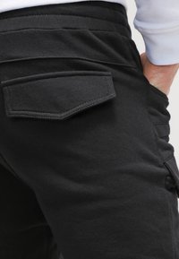 Černé bavlněné kalhoty s zadní kapsou obdélníkového tvaru a šitými okraji; hladká textura s mírně pružným pasem.