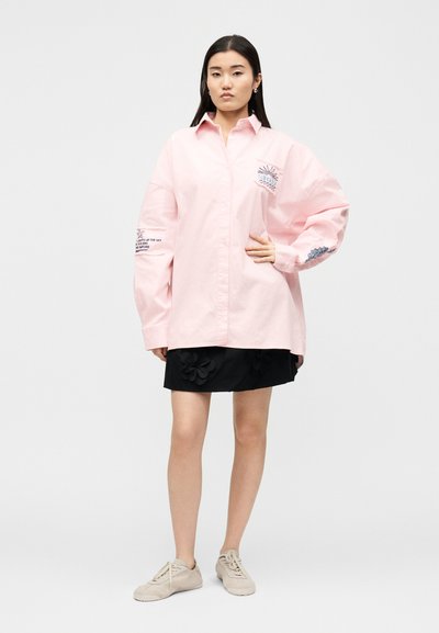 Γυναίκα στέκεται φορώντας υπερμεγέθη ανοιχτό ροζ πουκάμισο με τυπωμένα σχέδια στα μανίκια και το στήθος, μαύρη φούστα με λουλουδάτες λεπτομέρειες, μπεζ αθλητικά παπούτσια.