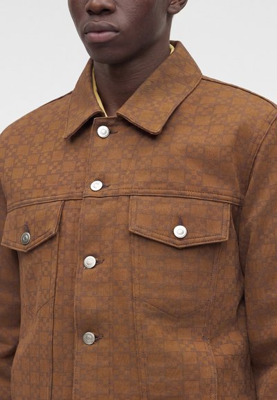 Homme portant une veste marron à carreaux avec des boutons argentés et des poches à rabats, montré de la poitrine vers le haut.