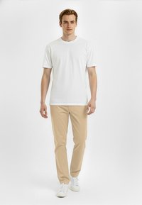 Hvid bomulds t-shirt med korte ærmer, parret med beige bukser og hvide sneakers, stående mod en ensfarvet baggrund.