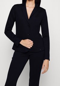 Blazer nero su misura con colletto a revers, chiusura con due bottoni, tasche frontali e design aderente, realizzato in un tessuto morbido.