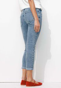 Jeans en denim bleu clair avec une coupe slim, ourlets retroussés et poches arrière, accompagnés de ballerines rouges. Le tissu semble texturé et légèrement délavé.