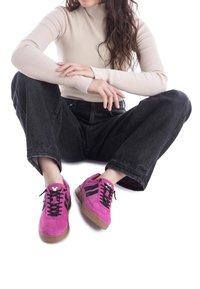 Zapatillas de ante rosa brillante con cordones negros, suelas de goma marrón y rayas negras. El modelo está sentado con vaqueros negros y una camiseta de punto beige.