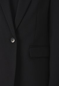 Musta bleiseri, jossa on yksi musta nappi ja läppätasku oikealla puolella, valmistettu sileästä kankaasta.