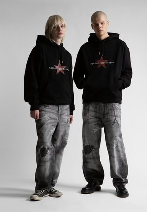 Δύο άτομα που φορούν μαύρες φούτερ με κουκούλα με κόκκινο αστέρι και το κείμενο "against perfection", φθαρμένα γκρι τζιν, στέκονται μπροστά σε απλό φόντο.