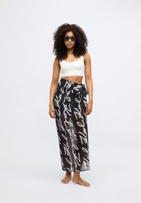 Sarong nero trasparente con motivo di testo bianco scritto a mano, legato in vita; abbinato a un crop top bianco aderente e occhiali da sole.