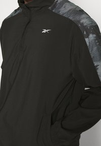Chaqueta ligera negra con media cremallera, con acentos grises abstractos en los hombros y un pequeño logo blanco en el pecho.