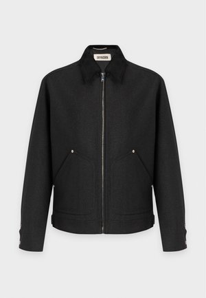 Homme portant un bonnet noir, une veste zippée noire, une chemise vert foncé, un pantalon noir et des chaussures habillées noires, debout sur fond blanc.