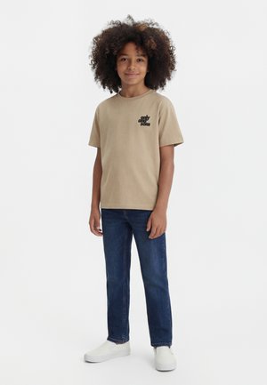 Niño con cabello rizado que lleva una camiseta beige con texto negro, vaqueros azules y zapatos blancos sin cordones, de pie sobre un fondo blanco.