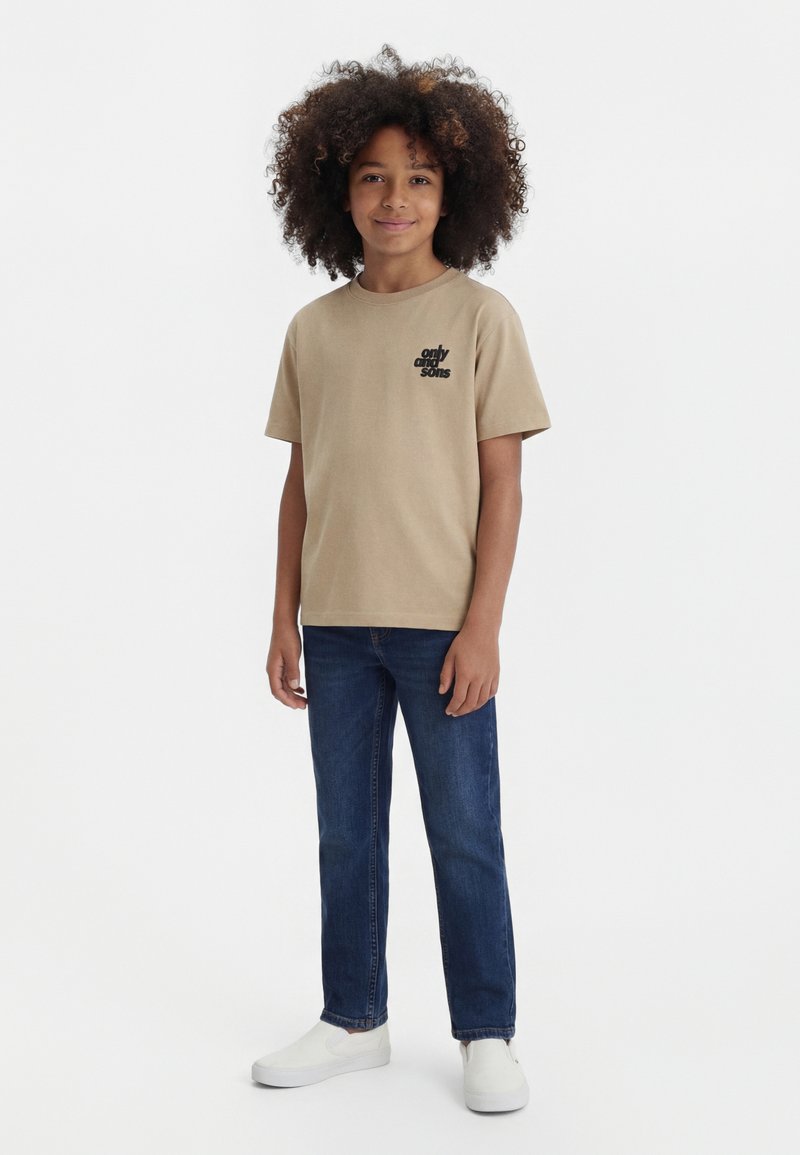 Bambino con capelli ricci che indossa una maglietta beige con scritta nera, jeans blu e scarpe slip-on bianche, in piedi su sfondo bianco.