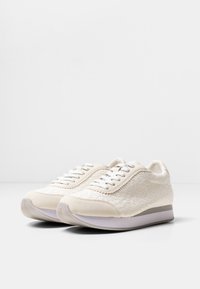 Zapatillas beige con tejido texturizado, detalles en cuero suave, punta redonda y cordones blancos. Suela de goma de color claro con un patrón de surcos sutil.