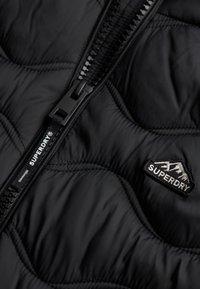 Zwarte gewatteerde pufferjas met een centrale rits. Voorzien van een zwarte ritsen trekker en een prominent "Superdry" logo patch met bergafbeeldingen.