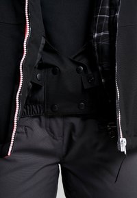 Schwarze Jacke mit Reißverschluss und Druckknopf-Verschlüssen, ausgestattet mit einem weichen Innenfutter, und schwarze Hosen mit glatter Textur und Taschen.