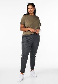 T-shirt a maniche corte verde oliva abbinato a pantaloni tapered grigi scuri, con un taglio slanciato e sneakers bianche. Outfit semplice e casual.