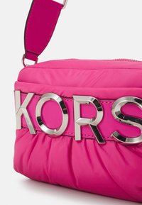 Różowa torebka crossbody z nylonu o marszczonej fakturze, z błyszczącym srebrnym logo "KORS" na kontrastującym pasku. Zamek błyskawiczny u góry.