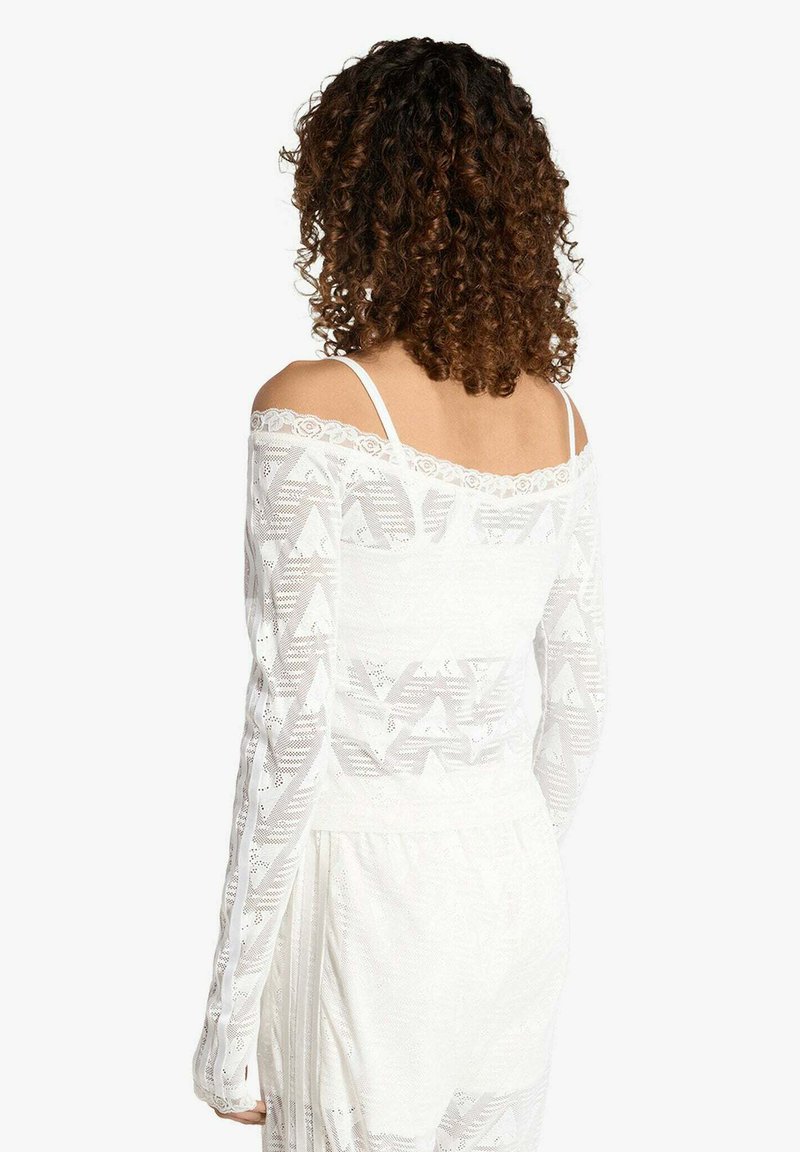 Lockige Frau von hinten gesehen, trägt ein weißes, schulterfreies Spitzenoberteil mit geometrischen Mustern und passende weiße Unterteile.