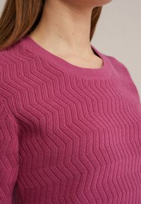 Detail osoby nosící texturovaný pletený svetr v barvě prachové růže se vzorem cikcak žebrování a zaobleným výstřihem.