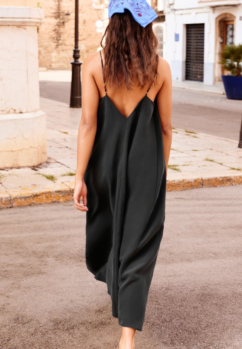 Femme aux cheveux ondulés portant une robe noire ample et dos nu avec un foulard bleu sur la tête, marchant dans une rue pavée calme.