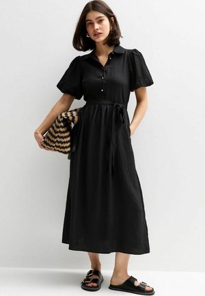 Black, knee-length dress with a buttoned front, short puff sleeves, and a tie belt. Paired with black sandals and a striped bag.