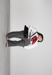 Γυναίκα με γκρι παντελόνι, λευκό πουκάμισο με κόκκινο μαντήλι σε σχήμα καρδιάς, μαύρες σαγιονάρες, κρατώντας μαύρη τσάντα, στέκεται μπροστά σε μονόχρωμο γκρι φόντο.