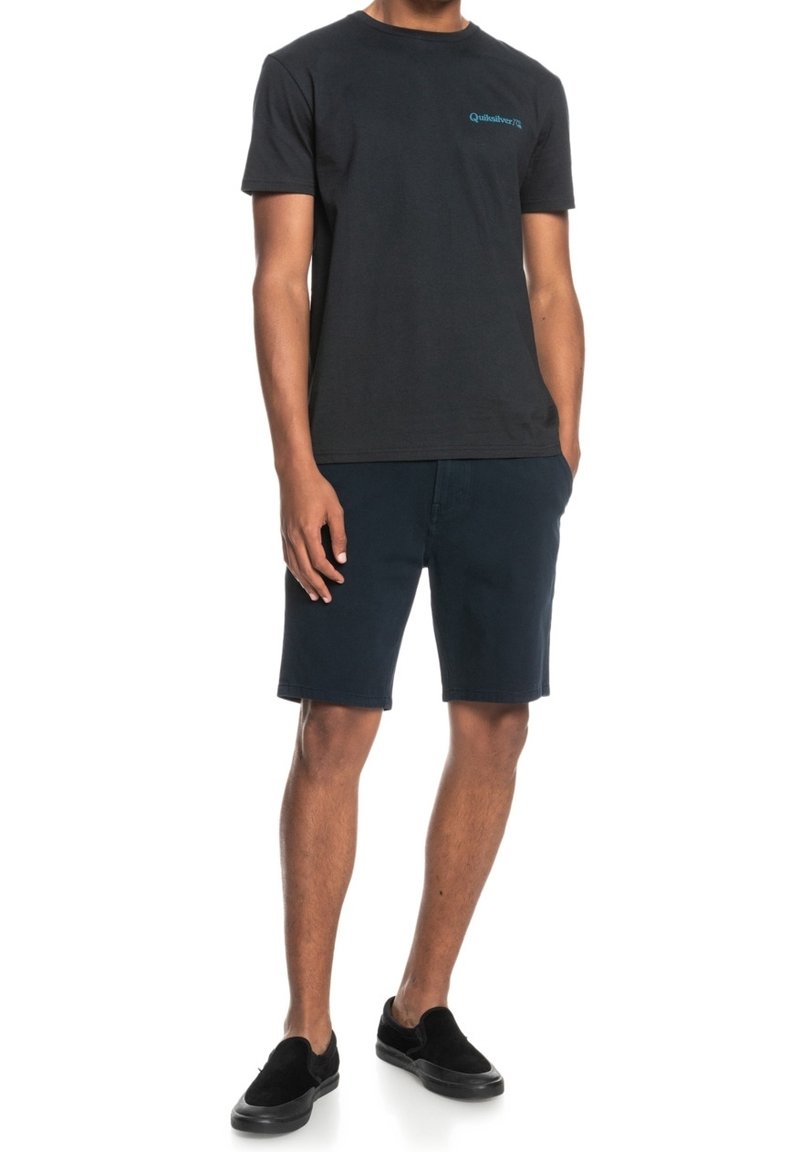 Homme portant un t-shirt noir Quiksilver, un short bleu marine foncé et des chaussures noires sans lacets, debout les mains dans les poches devant un fond blanc.