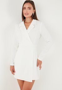 Woman wearing a white long-sleeve blazer dress with lapel collar, standing against a plain white background, with earrings and bracelet.