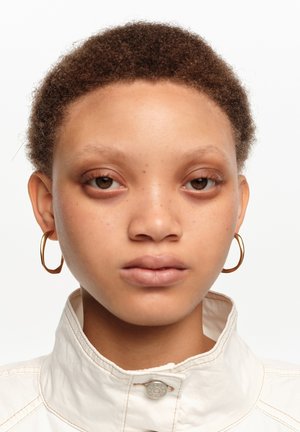 Joven con cabello corto y rizado, lleva pendientes de aro dorados y una chaqueta blanca de cuello alto, mirando directamente al frente.