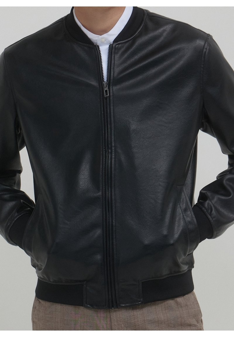 Homme portant une veste bomber en cuir noir zippée, avec poignets et taille côtelés, les mains dans les poches sur un pantalon marron clair.