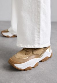 Beige mesh and leather sneaker with a thick white and brown rubber sole, featuring round toe and textile accents.