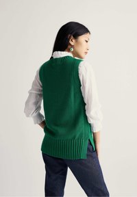 Chaleco verde de punto con dobladillo acanalado y aberturas laterales, llevado sobre una blusa blanca con puños con volantes, combinado con pantalones de mezclilla oscuros.