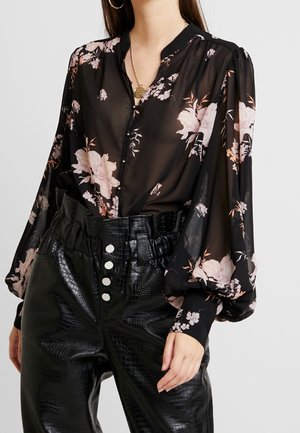 Blouse noire transparente à imprimé floral rose avec manches bouffantes, associée à un pantalon taille haute en simili cuir noir avec des boutons blancs.