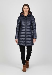 Marineblå pufferjakke med hætte, quiltet design, blank overflade og lynlås foran. Båret over sorte bukser og brune ankelstøvler.