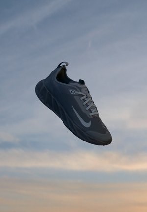 Zapatilla deportiva gris oscuro con logo y cordones blancos flotando contra un fondo de cielo nublado al atardecer.