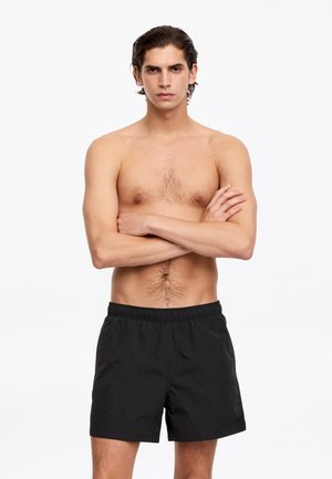 Ung mand med mellemlangt mørkt hår står barbent med armene over kors iført sorte shorts mod en hvid baggrund.