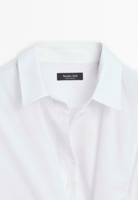 Biała bawełniana koszula z klasycznym kołnierzykiem, o gładkiej fakturze i przeszytym dekolcie. Metka głosi "Massimo Dutti, wyprodukowano w Maroku."