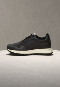 Μαύρο αθλητικό παπούτσι χαμηλού ύψους με λευκή σόλα, υφασμένο πέλμα, δέσιμο με κορδόνια μπροστά και μικρό μεταλλικό λογότυπο στο πλάι, σε ουδέτερο φόντο.