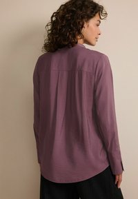 Langærmet, afslappet skjorte i mauve med rygplissé-detalje, subtil tekstur og buet kant, parret med mørke stribede bukser.
