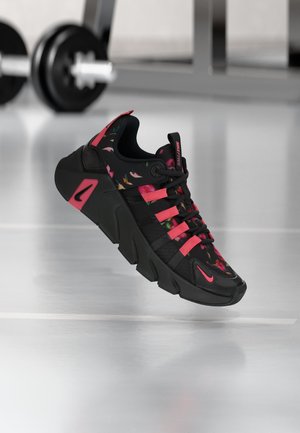 Baskets Nike Free noires et roses avec des détails floraux, une semelle épaisse et des lacets, présentées dans une salle de sport avec des poids en arrière-plan.
